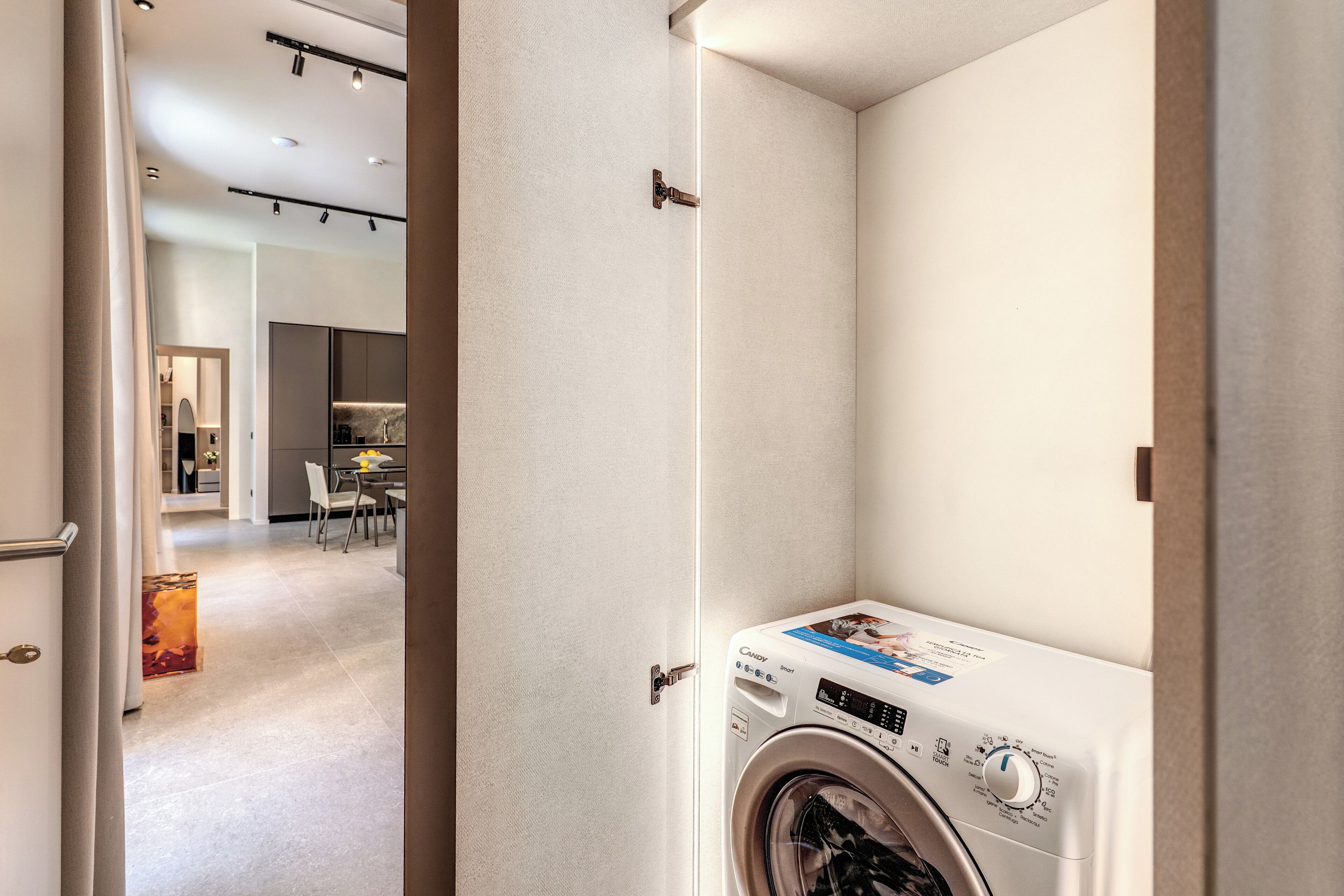 Une petite buanderie équipée d’un lave-linge à chargement frontal, avec une porte donnant sur un coin repas et une cuisine visible au fond.