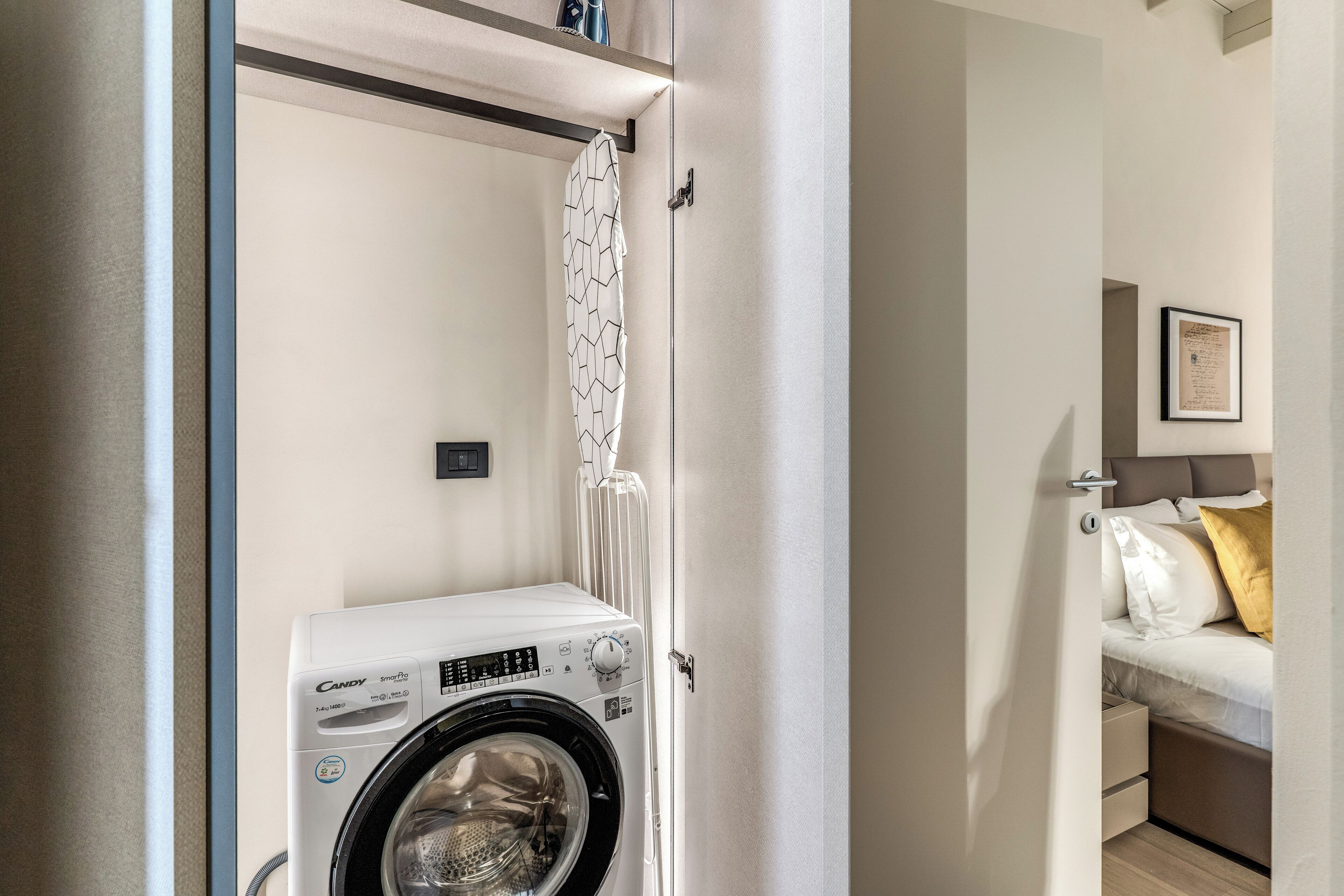 A compact laundry area with a Candy brand washing machine, a towel rack, and a bed with a headboard in the adjacent room.