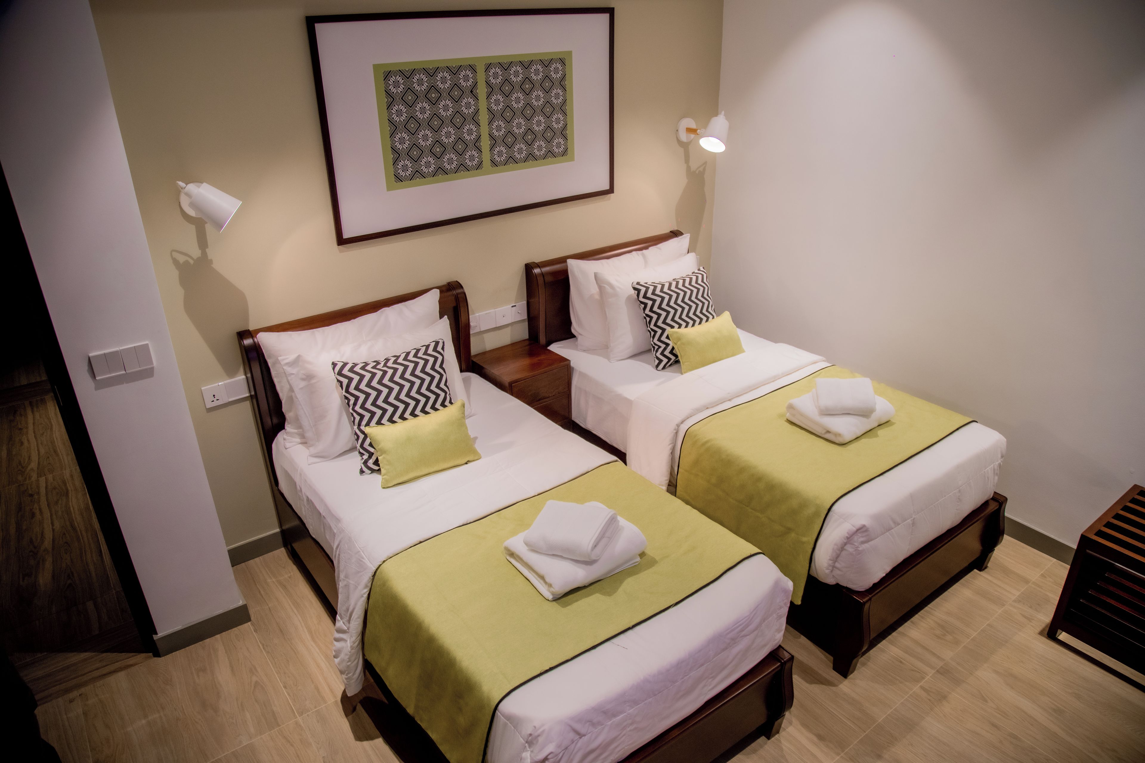 A hotel room with two beds, each with a green bedspread and white bedding. There are decorative pillows and a framed artwork above the beds.