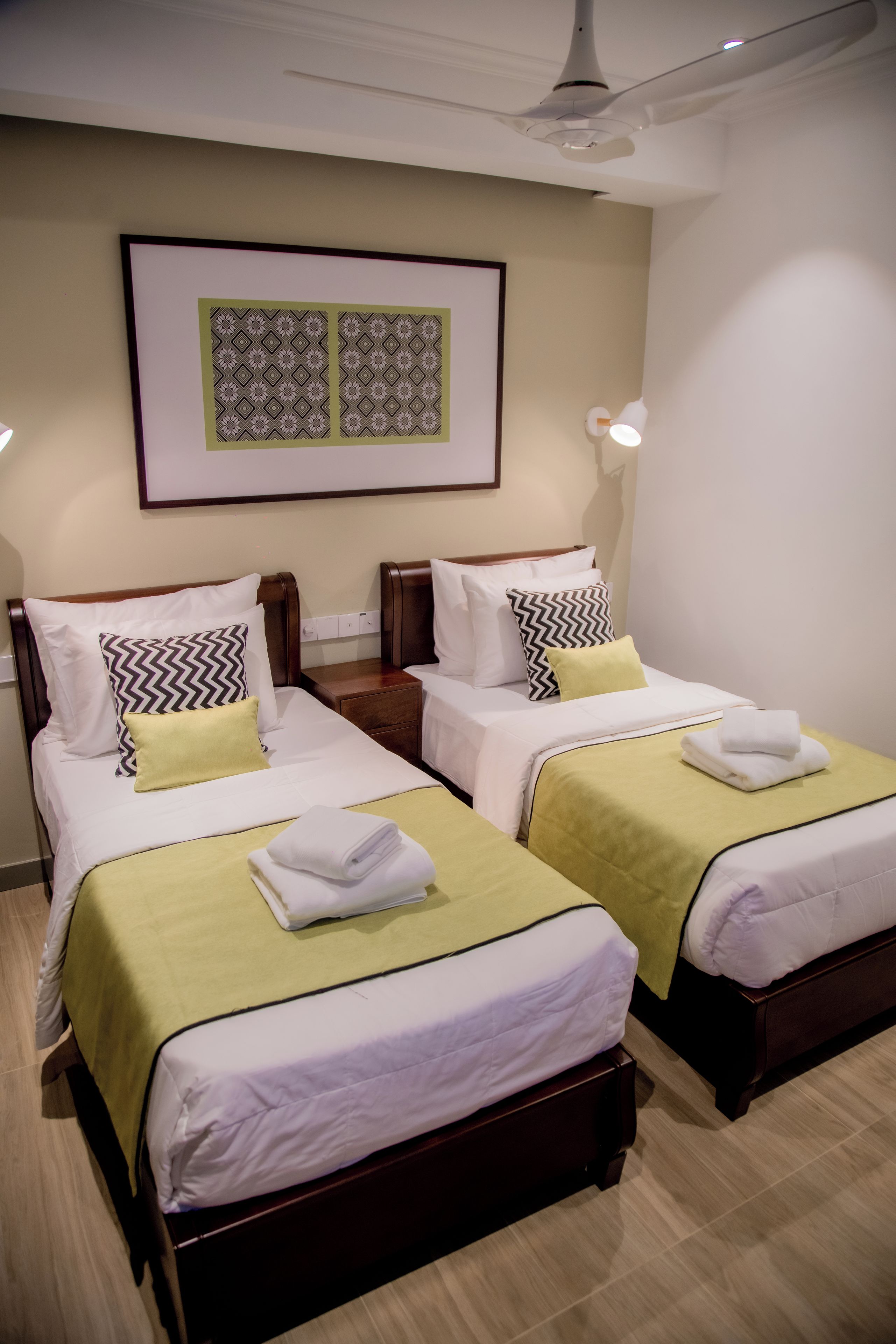 Une chambre d’hôtel avec deux lits, chacun recouvert d’une couverture verte et doté de draps blancs, une œuvre d’art encadrée au-dessus des lits et un ventilateur de plafond.
