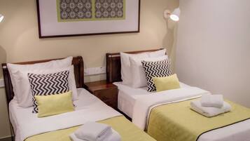 A hotel room with two beds, each with a green bedspread and white linens, a framed artwork above the beds, and a ceiling fan.