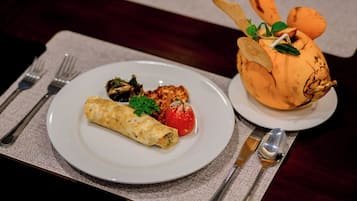 A plate with a rolled omelette, a grilled chicken breast, and a grilled tomato, accompanied by a bread roll with a decorative topping.