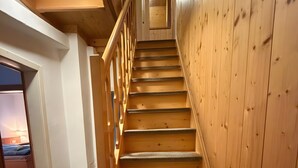 Una escalera de madera con barandilla, escalones alfombrados y una puerta al final que da a una habitación con cama y ventana.