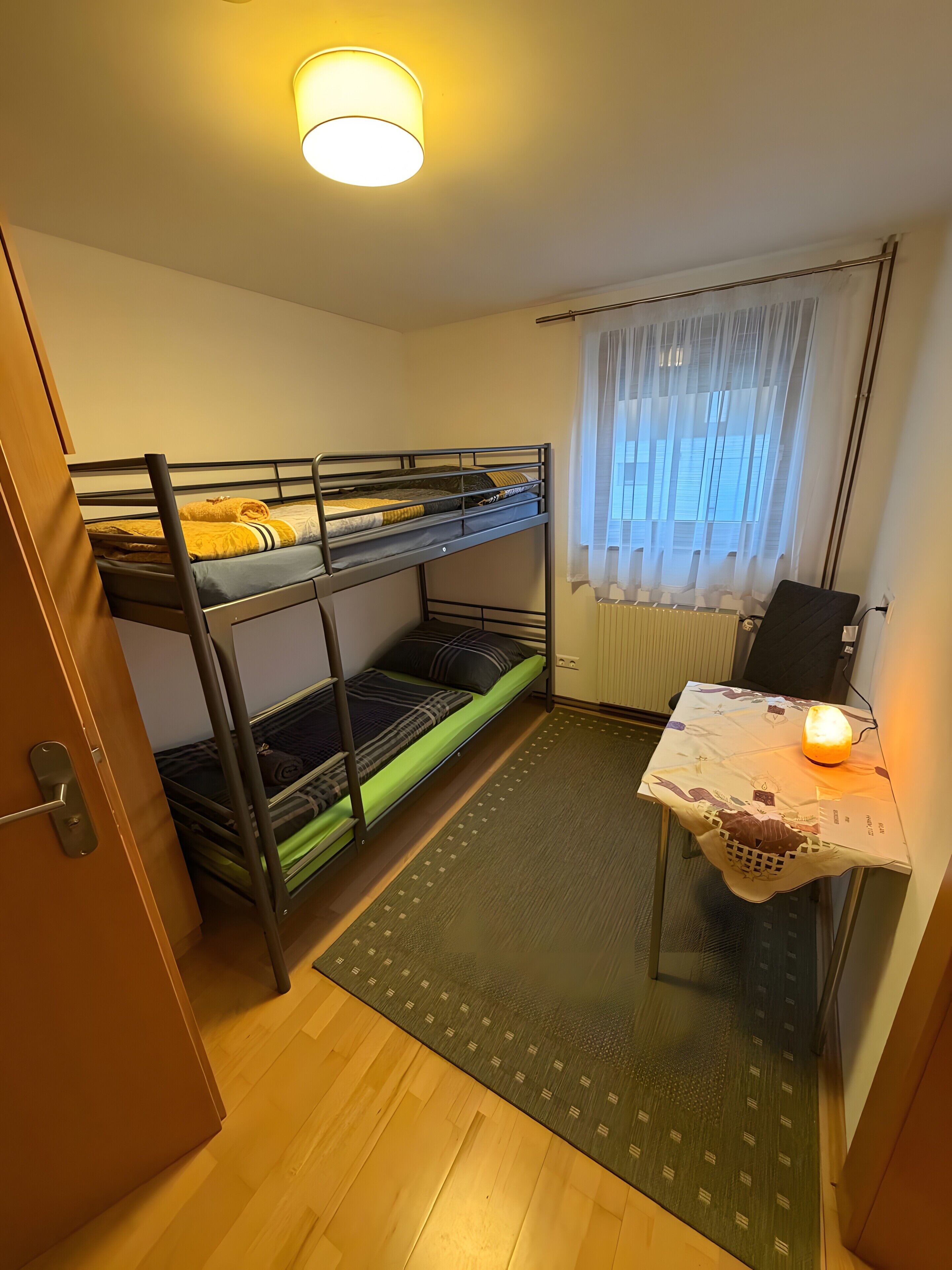 Ett litet rum med våningssängar, ett litet bord med en lampa och ett fönster med tunna, genomskinliga gardiner.