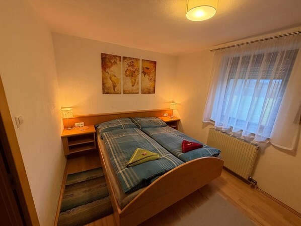 A bedroom with a bed, two bedside tables, a lamp, a world map artwork, a window with curtains, and a radiator.