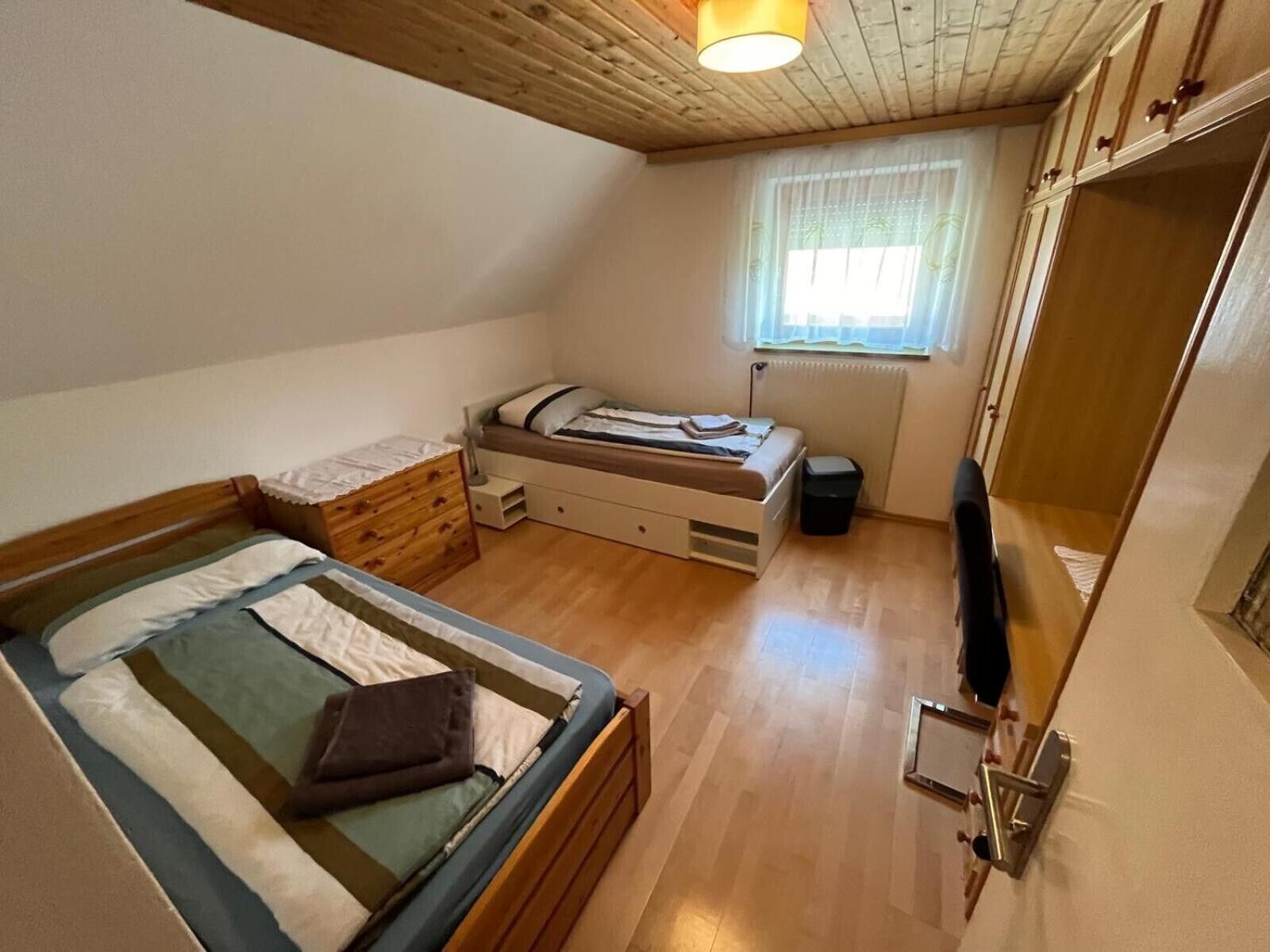 A small, cozy room with a bunk bed, a single bed, a window with curtains, and a wooden ceiling.