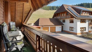 Un balcon en bois offrant une vue sur une maison équipée de panneaux solaires, un espace salon couvert et une terrasse délimitée par une clôture.