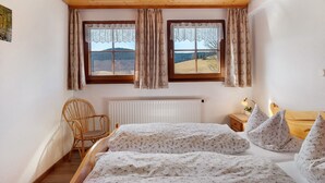 Une chambre avec un plafond en bois, deux fenêtres avec des rideaux, un radiateur, une table de chevet en bois avec une lampe et un fauteuil en osier.