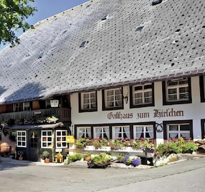 Um edifício tradicional com uma placa indicando 'Gasthof zum Hirschchen'. Possui telhado de telhas, várias janelas e um jardim frontal com vasos de plantas.