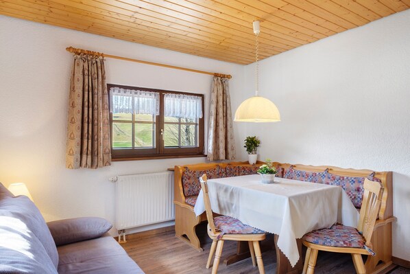 A cozy room with a wooden ceiling, a table with a white tablecloth, a bench with patterned cushions, and a window with curtains. - Talblick Apartment – Restaurant Open Mondays (Titisee-Neustadt)