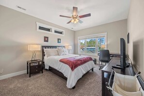 Une chambre à coucher comprenant un lit, des tables de chevet, un bureau avec un ordinateur, un ventilateur de plafond et une fenêtre donnant sur un beau paysage.