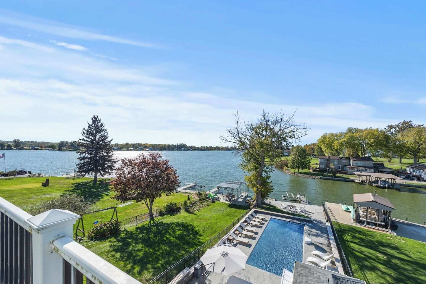 A pool with lounge chairs, a dock with boats, and a view of a lake with houses in the background.