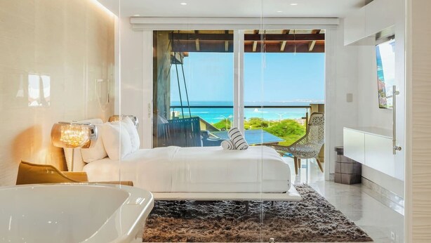 A modern bedroom with a large bed, a glass-enclosed balcony, a view of the ocean, and a bathroom with a bathtub. - 2 Mins to Beach w/ Ocean & Pool Views by Bocobay (Noord)