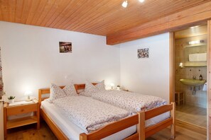 Ein hölzernes Bett mit einer blumigen Tagesdecke, ein Nachttisch mit Lampe sowie ein Badezimmer mit Waschbecken und Spiegel.