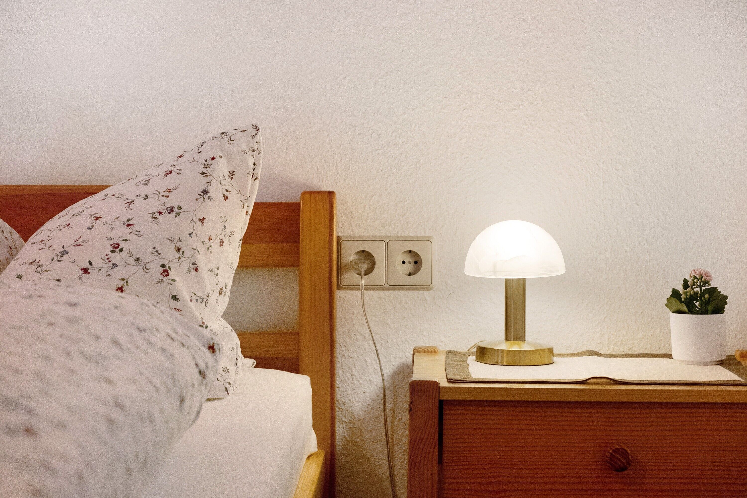 Un letto rifatto con biancheria da letto floreale, una lampada da comodino, un piccolo comodino in legno e una pianta in vaso.