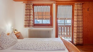 Quarto com teto de madeira, cama com roupa de cama floral, escrivaninha com abajur e janela com cortinas estampadas.