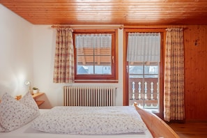Quarto com teto de madeira, cama com roupa de cama floral, escrivaninha com abajur e janela com cortinas estampadas.