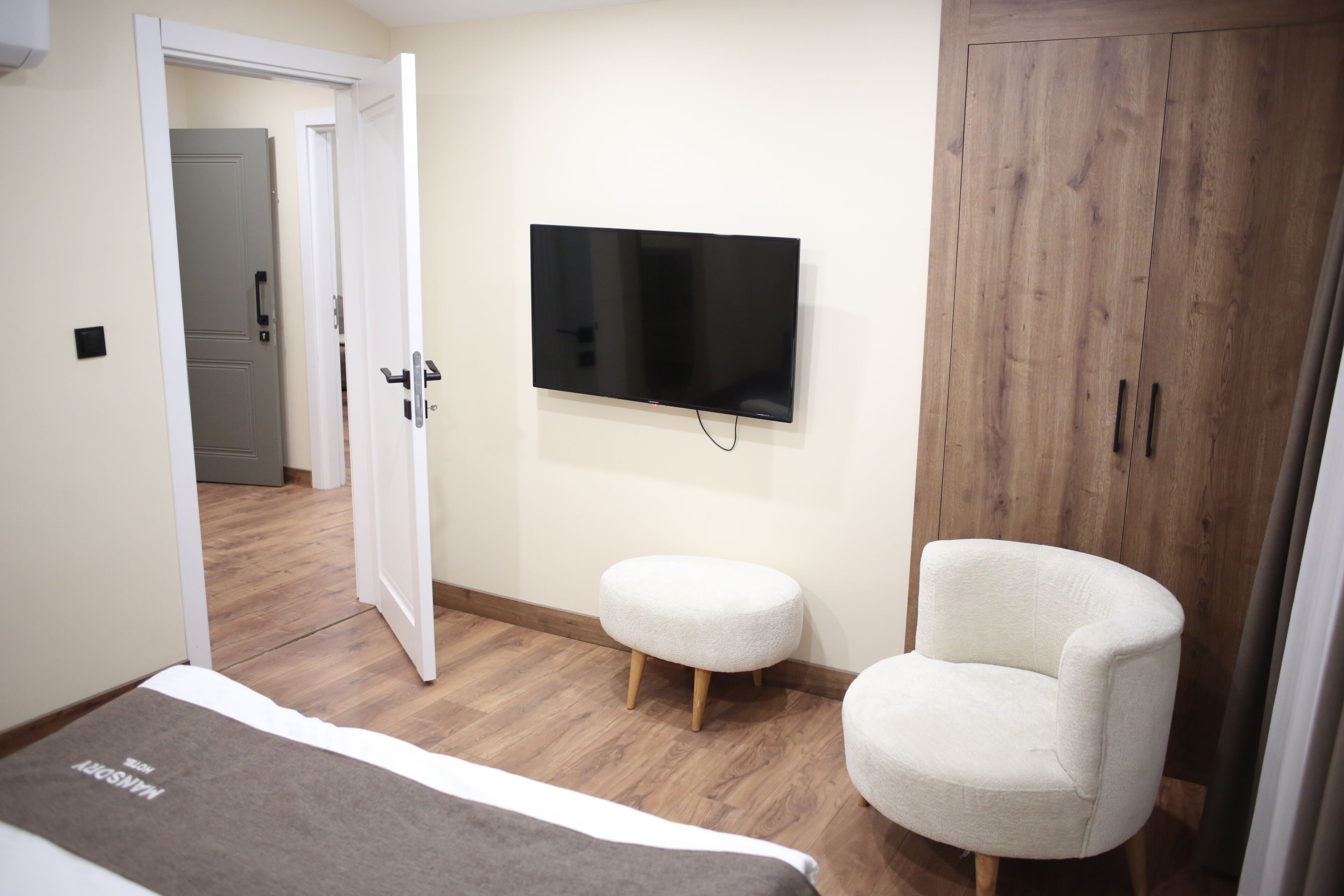 Un dormitorio con una cama, un televisor montado en la pared, un armario de madera y un sillón blanco.