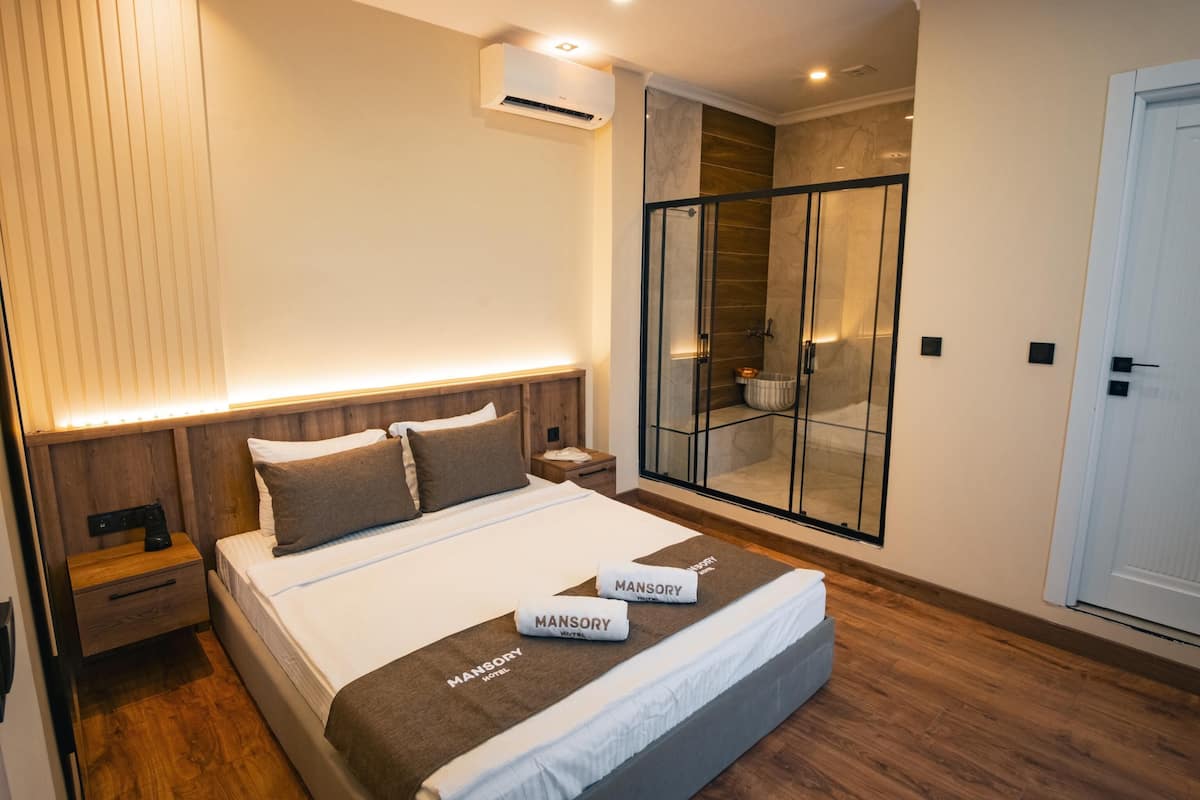 A modern hotel room with a large bed, wooden headboard, and a glass-enclosed shower area.