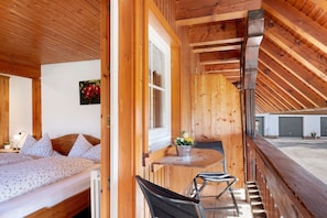 Een houten kamer met een bed, een raam, een tafel met stoelen en uitzicht op een gebouw.