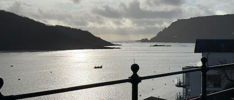 Vista costera con barandilla en primer plano, un barco en el agua y colinas al fondo.