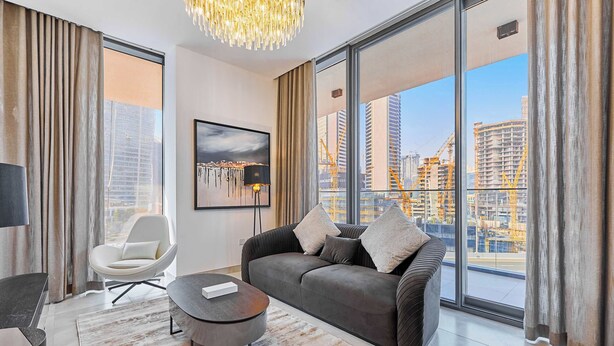 Een moderne woonkamer met een grijze bank, een witte fauteuil en een ronde salontafel. Een groot raam biedt uitzicht op een stadsgezicht met wolkenkrabbers en hoogwerkers.