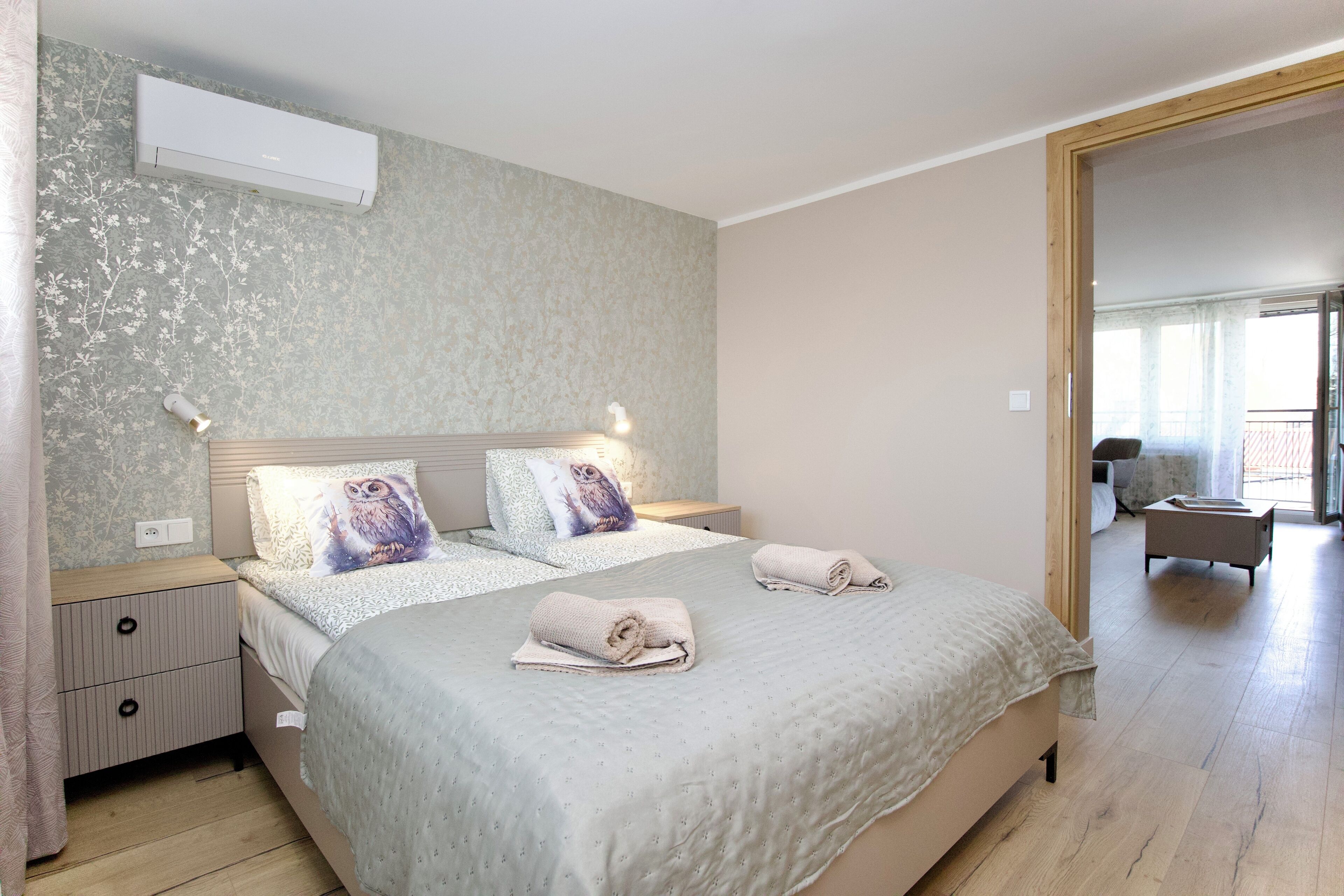 A bedroom with a bed, bedside tables, an air conditioner, and a view to another room.