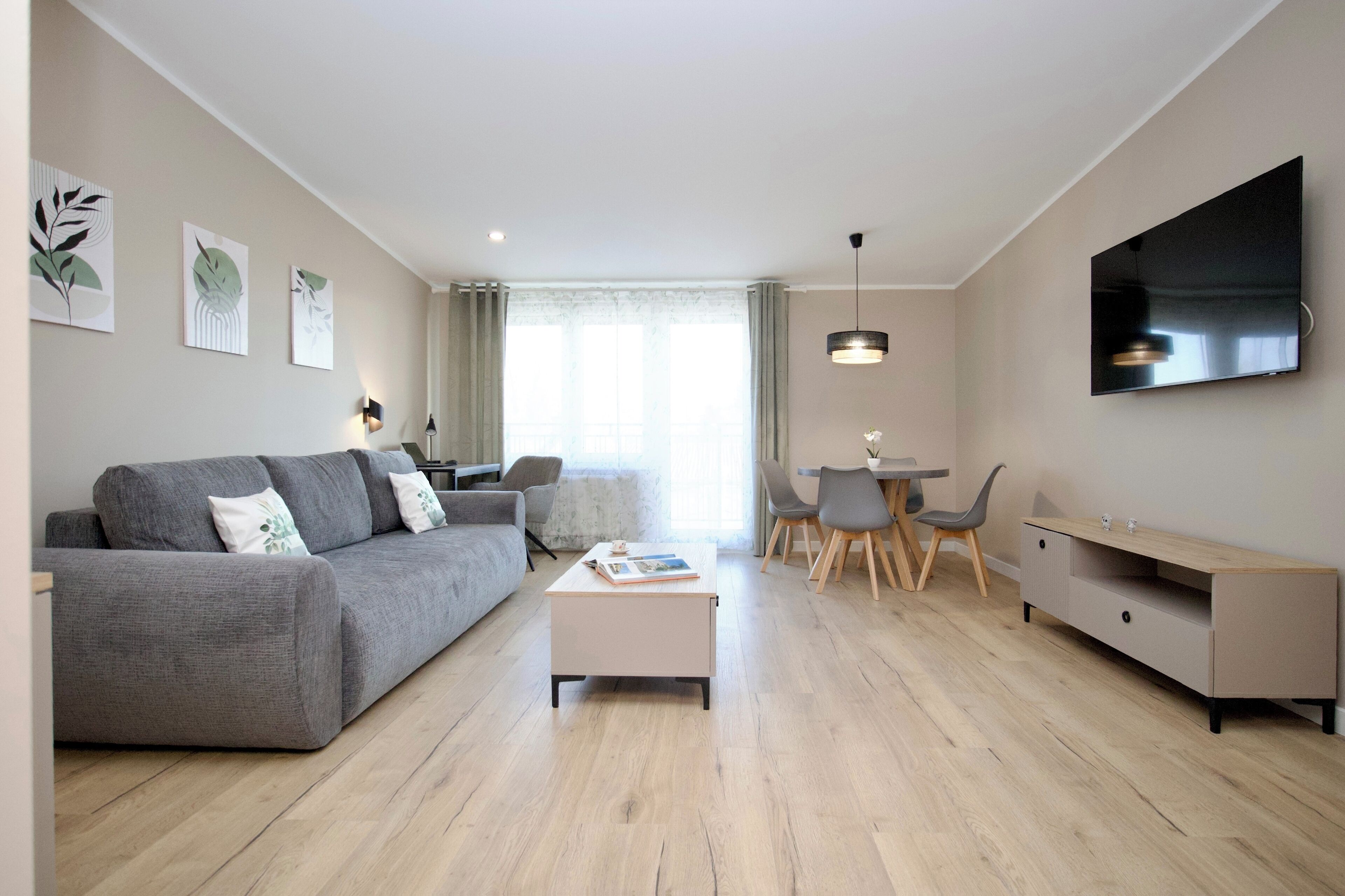 A modern living room with a grey sofa, a coffee table, a dining area with chairs, and a TV mounted on the wall.