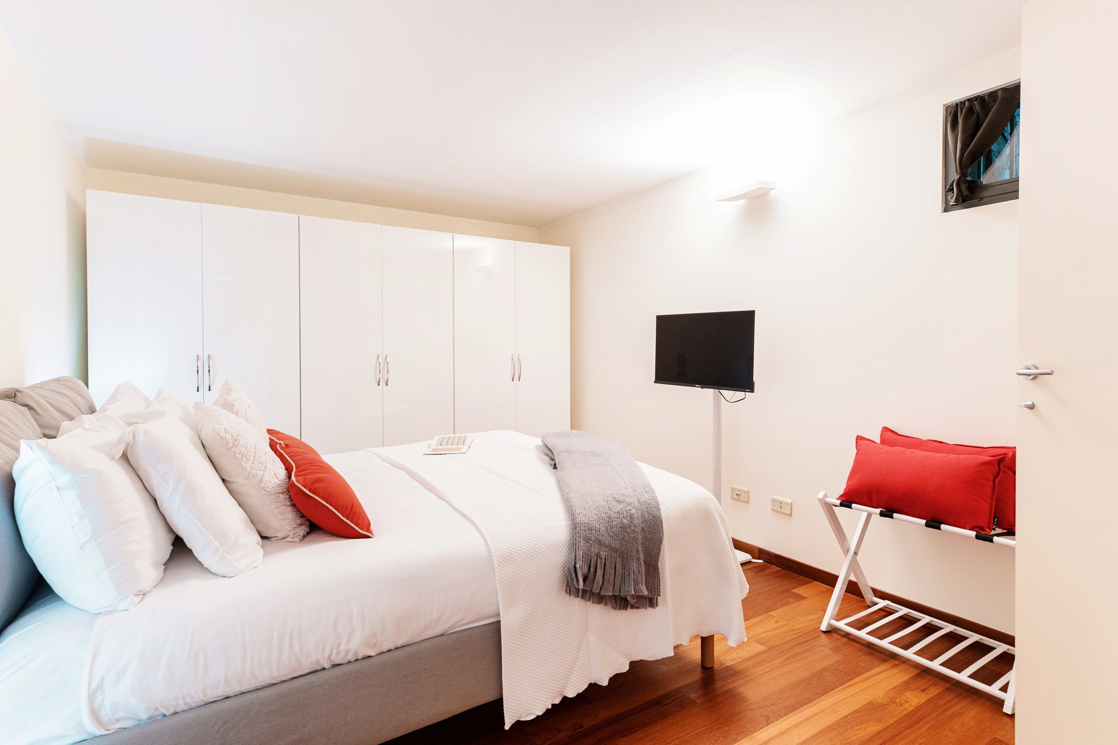 Ein Schlafzimmer mit Bett, weißem Kleiderschrank, Fernseher und einem roten Kissen auf einem Regal.