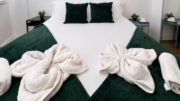 A neatly made bed with a green bedspread, two pillows, and two rolled towels.