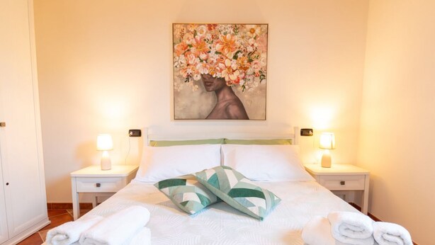 A neatly made bed with pillows, a painting above it, and two bedside lamps. - Domo Mea Rooms- Camera 1 (CABRAS)