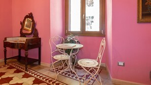 Een kamer met roze wanden, een houten bureau, een klein rond tafeltje met stoelen en een raam met kantgordijnen.