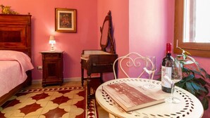 Een kamer met een bed, een klein tafeltje met een boek en wijnglazen, een nachtkastje met een lamp en een stoel.