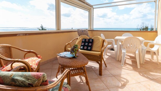 Un patio iluminado por el sol con sillas de mimbre, una mesita con cojín y vistas al mar.