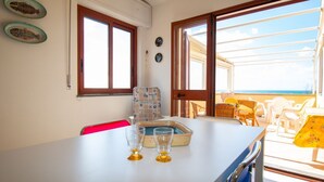 Una habitación con una mesa de comedor, dos sillas, una mesita con un plato y vasos, una ventana y vista al mar.
