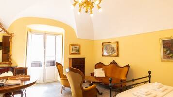 Una habitación con cama, sofá, mesa con libros, silla, candelabro y puerta que da al exterior.