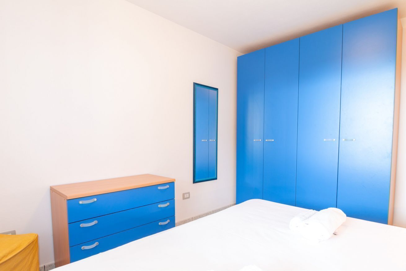 A bedroom with a bed, a blue wardrobe, a small dresser, and a wall-mounted mirror.