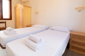 A hotel room with two single beds, each with a neatly folded white towel. There is a wooden wardrobe and a bedside table with a lamp.