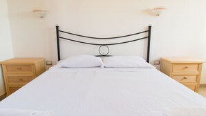 Een keurig opgemaakt bed met witte lakens, twee nachtkastjes met lades en wandlampen.
