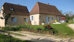 Une maison en pierre à deux étages, avec un toit en pente, un chemin de gravier menant à l’entrée et un petit jardin agrémenté de fleurs et d’une pelouse.