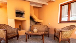 A cozy outdoor seating area with wicker furniture, a stone floor, and a fireplace. - S'Imbucada Home-Mare e Relax (SAN VERO MILIS)