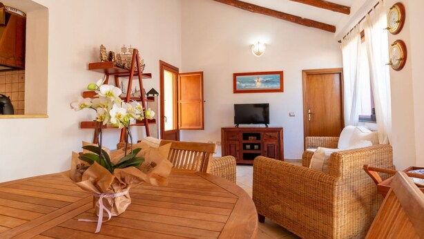 A cozy living room with a wooden dining table, wicker furniture, a TV, and a painting on the wall. - S'Imbucada Home-Mare e Relax (SAN VERO MILIS)