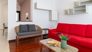 Un salon moderne avec un canapé en osier, un divan rouge, une table basse en verre sur laquelle repose un magazine, et une plante en pot.