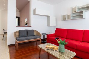 Una sala moderna con un sofá de mimbre, un sofá rojo, una mesa de centro de vidrio con una revista y una planta en maceta.
