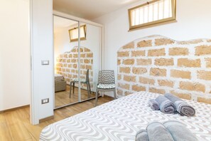 Une chambre à coucher avec une tête de lit ornée d’un motif en briques, un lit avec une literie à motifs, une chaise avec un coussin et un miroir qui reflète la pièce.