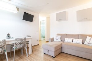 Una sala de estar moderna con un sofá, una mesa de comedor con sillas, un televisor montado en la pared y una pequeña cocina.