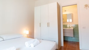 Une chambre d’hôtel avec un lit, une armoire, une table de chevet avec une lampe, et une salle de bain visible à travers une porte ouverte.