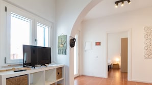 Un salon moderne doté d’un téléviseur, d’étagères de rangement, d’une grande fenêtre et d’un couloir menant à une chambre.