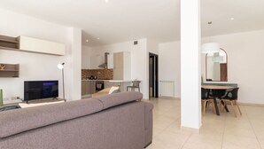 Un salon moderne avec un canapé, un téléviseur, un coin repas comprenant une table et des chaises, et une cuisine en arrière-plan.