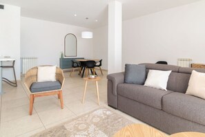 Un salon moderne comprenant un canapé, un fauteuil en osier, une table à manger avec des chaises et un miroir au mur.
